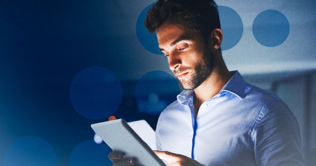 Homem de barba analisando tablet iluminado em fundo azul com bolhas bokeh, simbolizando operação de TI.