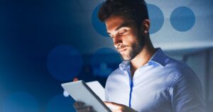 Homem de barba analisando tablet iluminado em fundo azul com bolhas bokeh, simbolizando operação de TI.